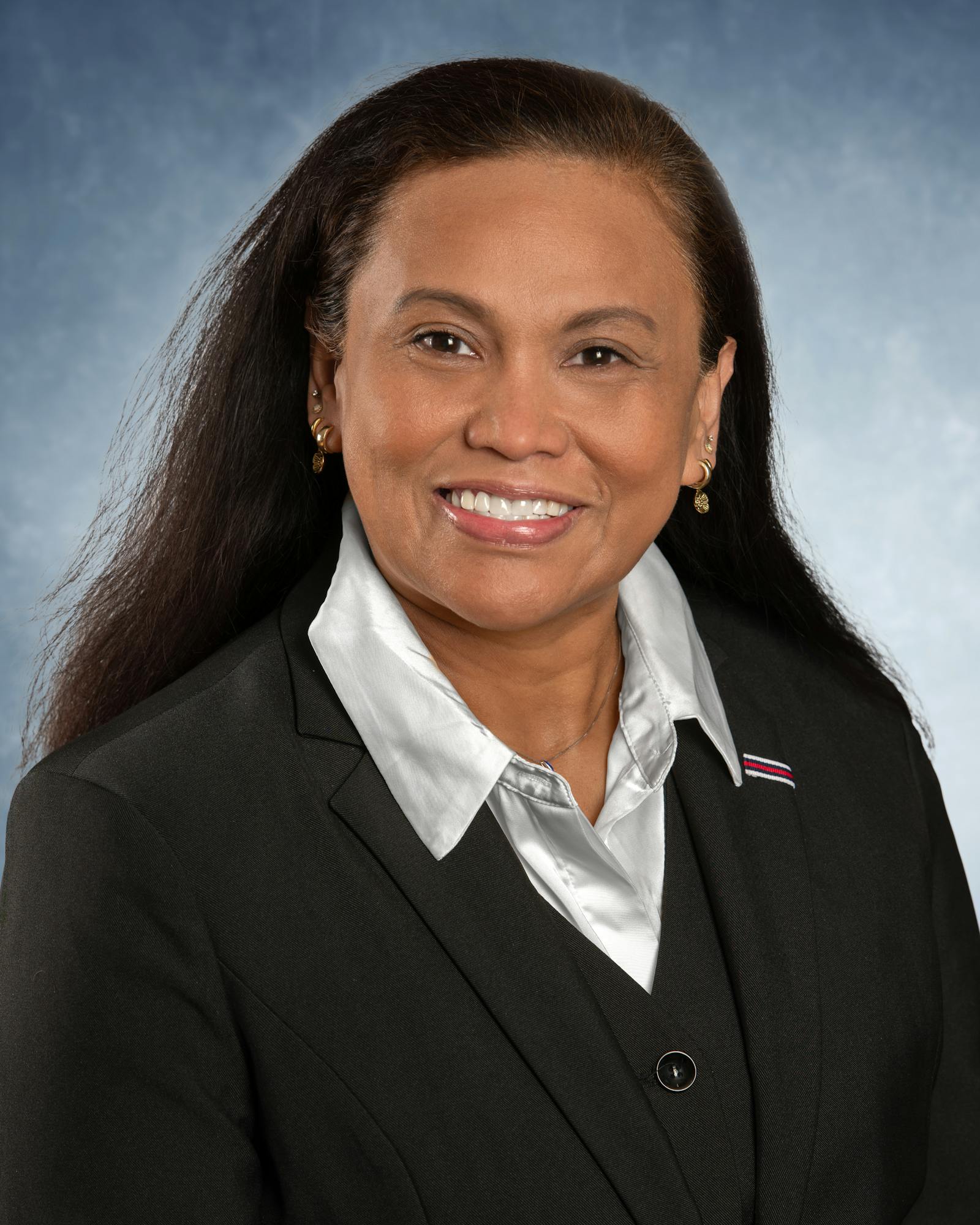 A person in a black suit and a white shirt, smiling against a blue background.