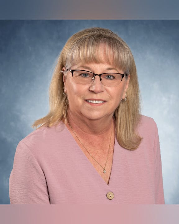 A woman with glasses and blonde hair, wearing a pink top, smiles against a blue background.