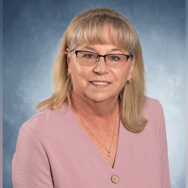 A woman with glasses and blonde hair, wearing a pink top, smiles against a blue background.
