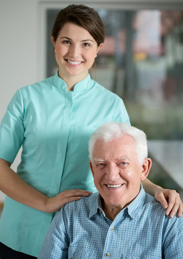 A smiling woman in a light blue shirt stands behind an elderly man in a plaid shirt, both looking at the camera.
