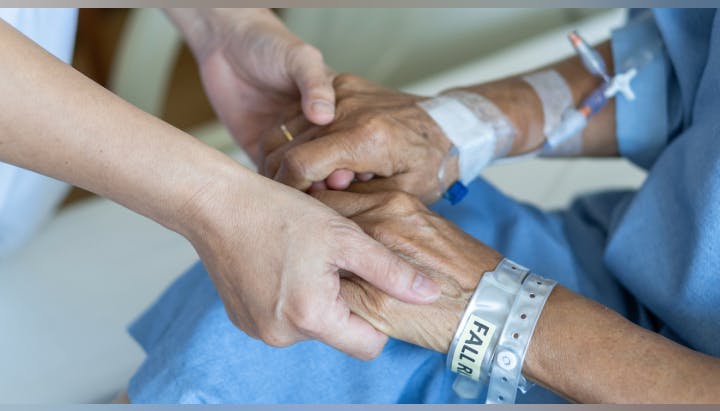 A caregiver holds the hands of an elderly patient with medical wristbands and IV, providing comfort and support.