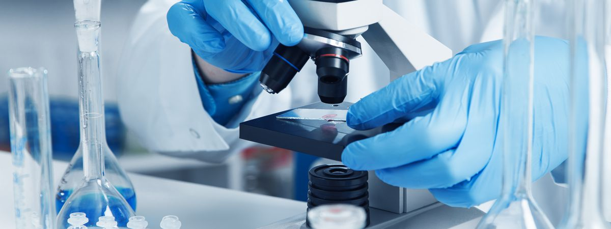 Hands in blue gloves using a microscope with lab equipment nearby.