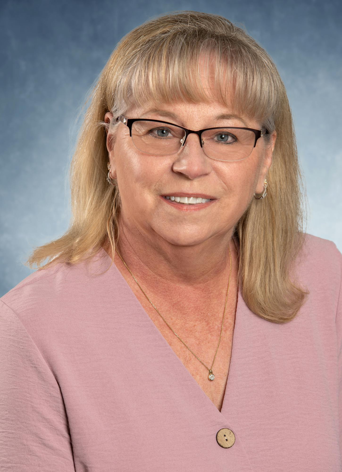 A woman with glasses and shoulder-length blonde hair, wearing a pink top, poses in front of a blue background.