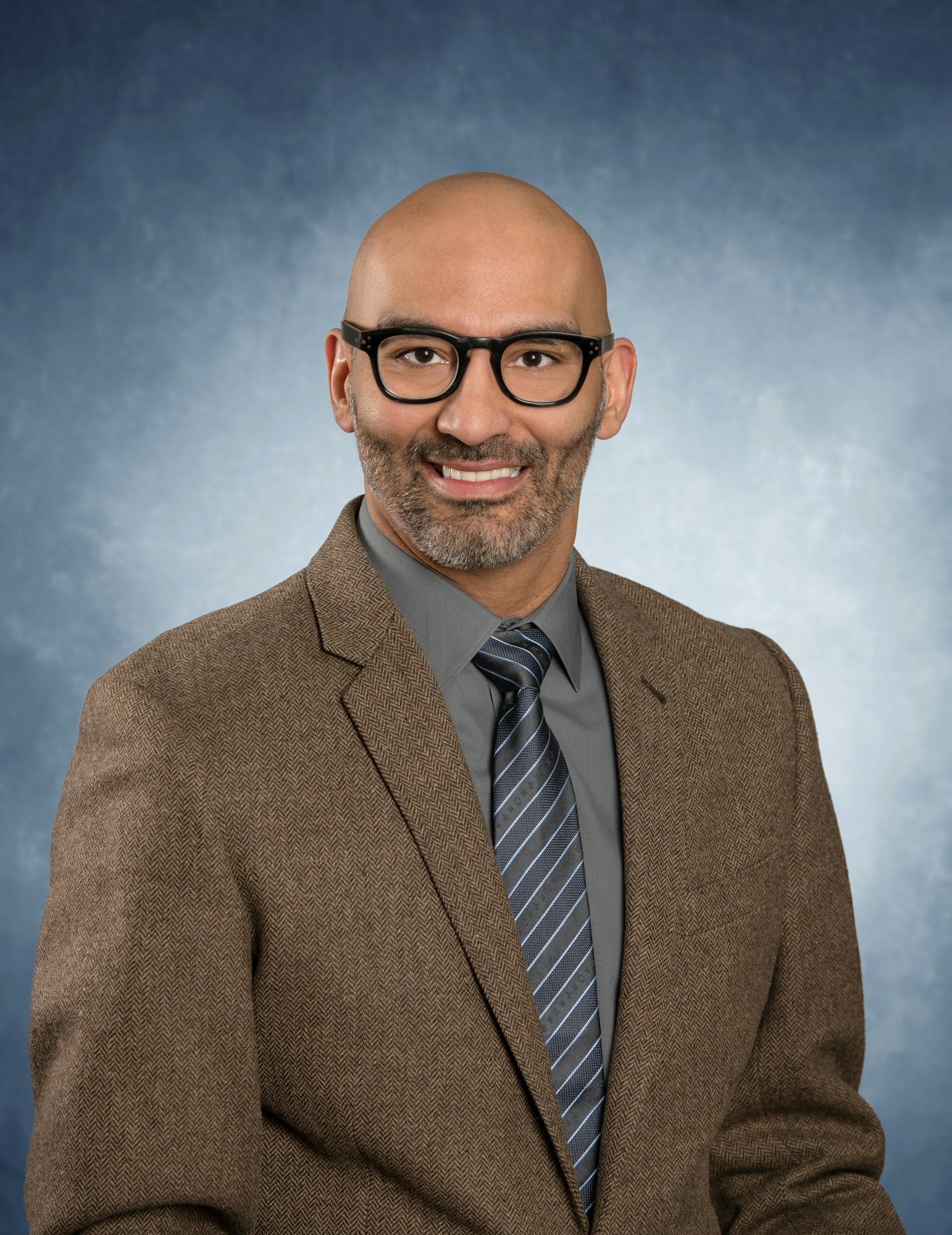 A person wearing glasses, a brown suit jacket, a gray shirt, and a striped tie, smiling against a textured blue background.