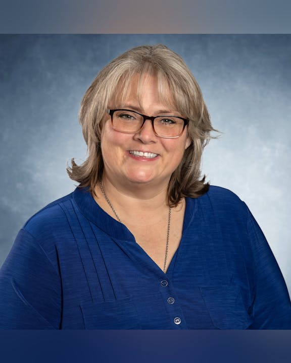 Smiling person with glasses, wearing a blue shirt against a blue-gray background.