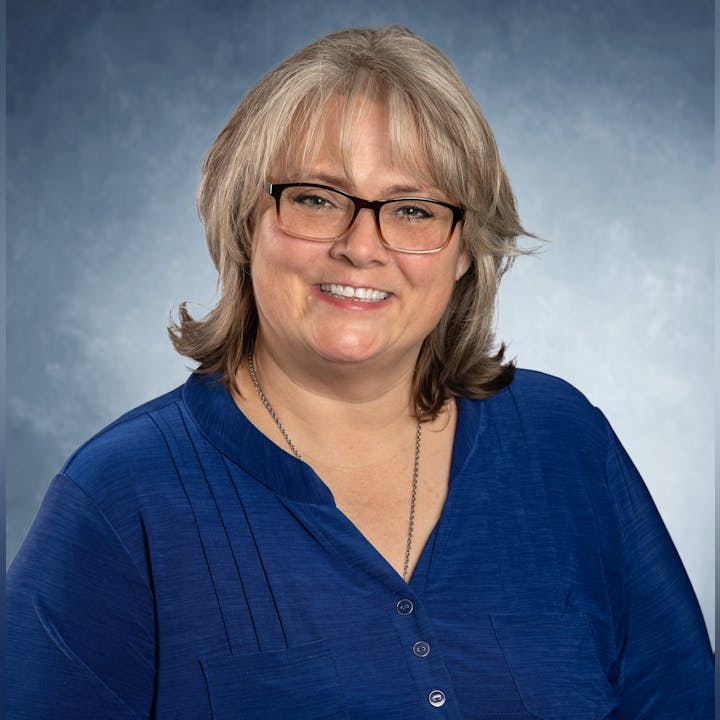 Smiling person with glasses, wearing a blue shirt against a blue-gray background.