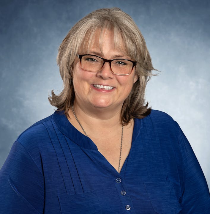 Smiling person with glasses, wearing a blue shirt against a blue-gray background.
