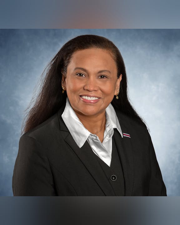 A person smiling, wearing a black suit and white shirt, against a blue background.