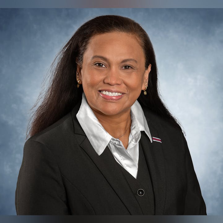 A person smiling, wearing a black suit and white shirt, against a blue background.