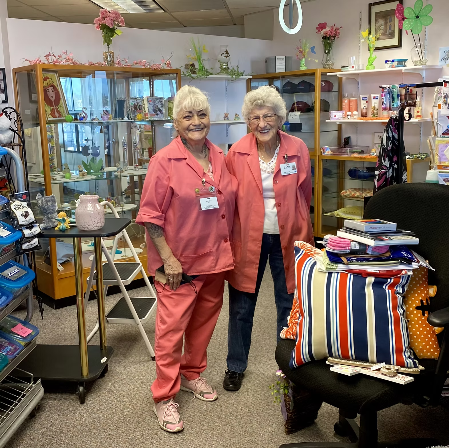 Two smiling women in matching pink outfits stand in a shop with shelves of various items.