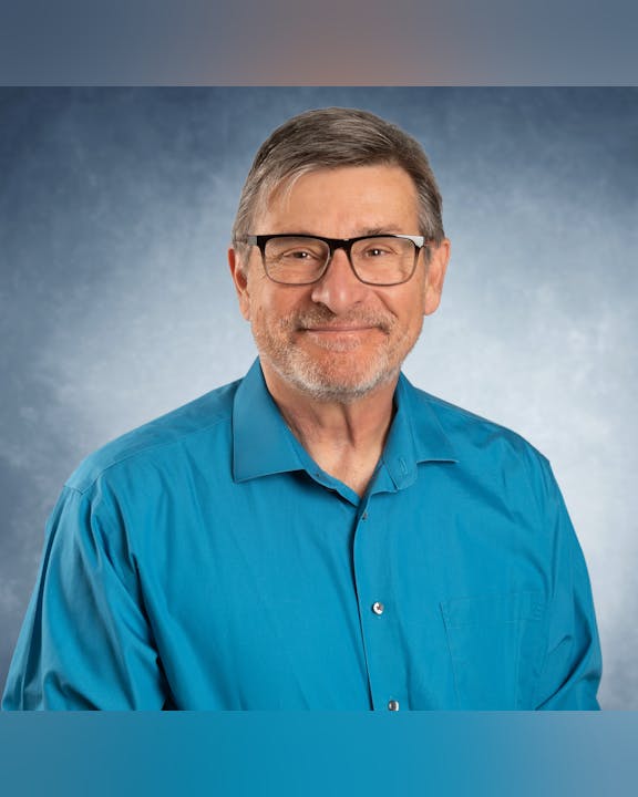 A man with glasses wearing a blue shirt, smiling against a gray background.