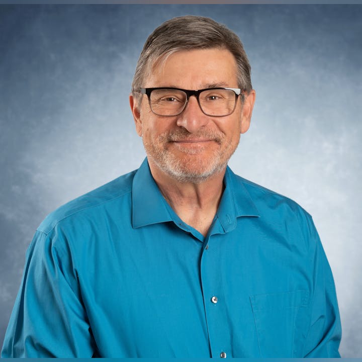 A man with glasses wearing a blue shirt, smiling against a gray background.