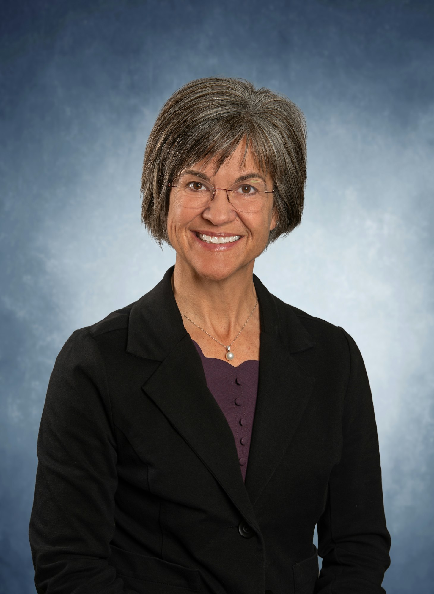 Person smiling, wearing glasses and a black jacket, with a blue textured background.