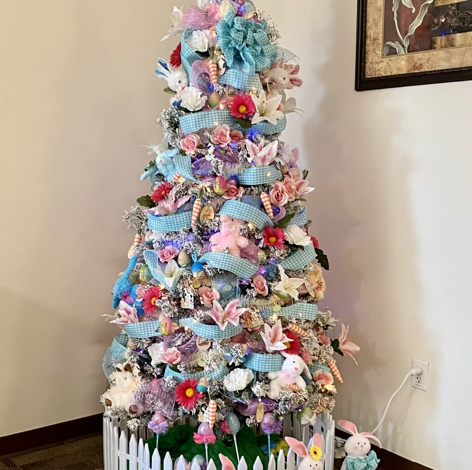 A festive tree adorned with pastel flowers, ribbons, Easter eggs, and bunnies in a white picket fence.