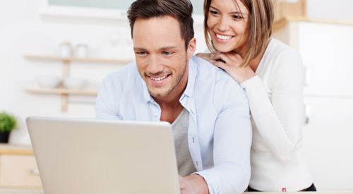 A man and woman smiling while looking at a laptop together in a bright room.