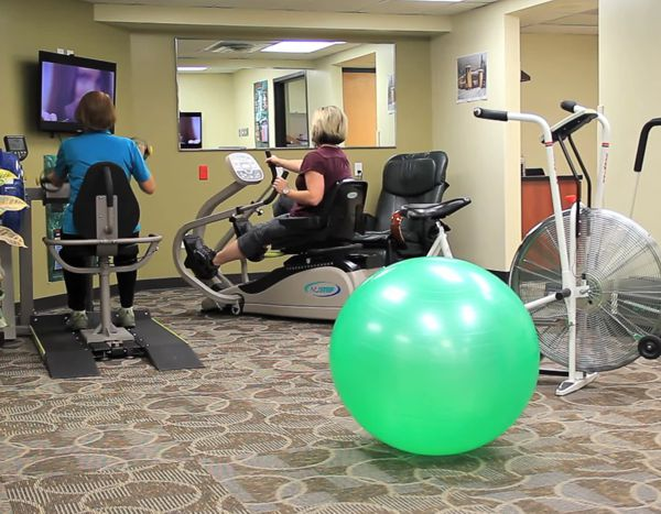 Two people on exercise bikes, a green exercise ball, and a fan bike in a gym setting.
