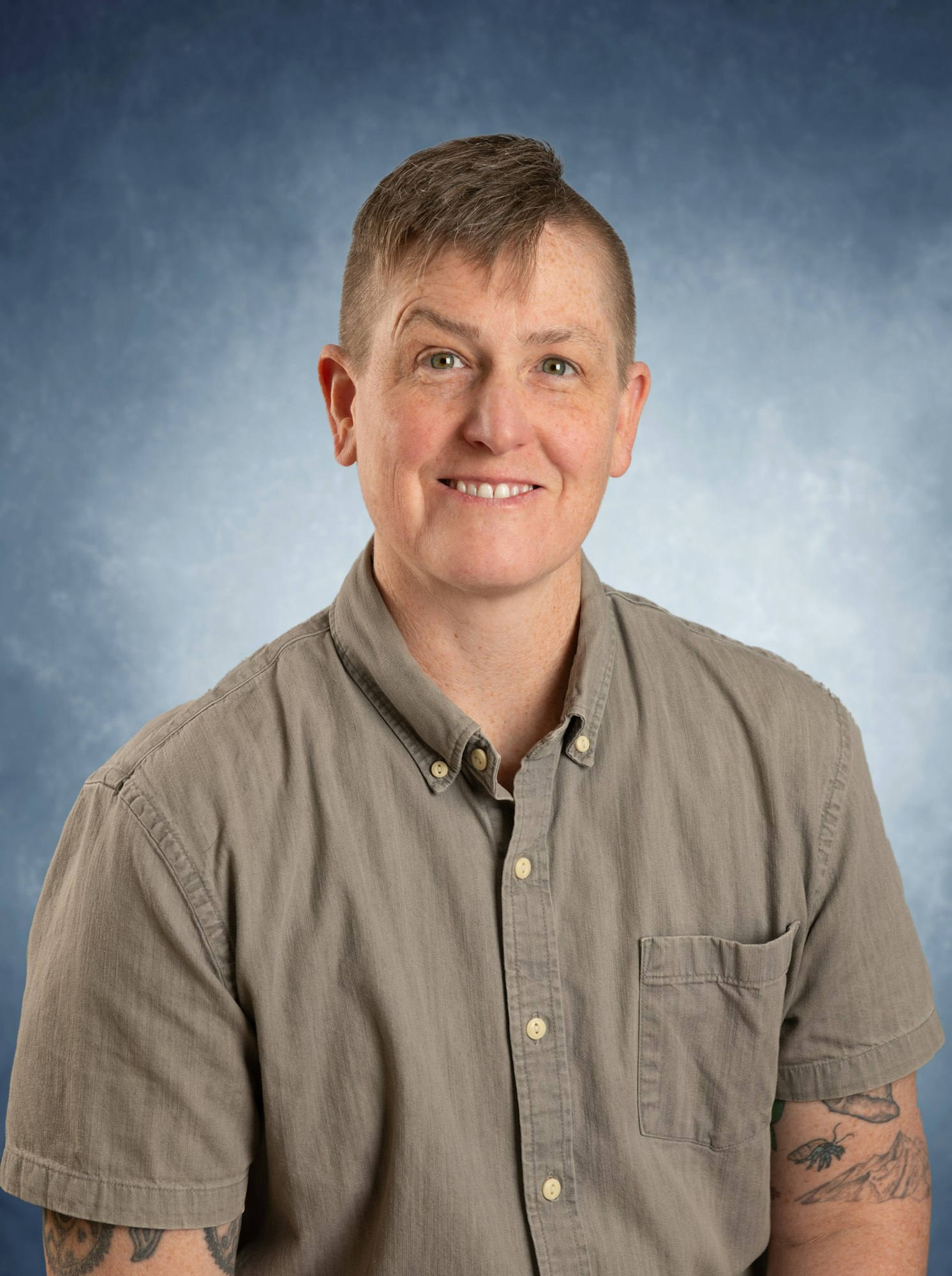 A person with short hair wearing a gray button-up shirt, smiling against a blue background.