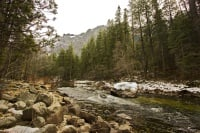 A river flows through a rocky bed with snow patches and trees against a backdrop of foggy mountains.