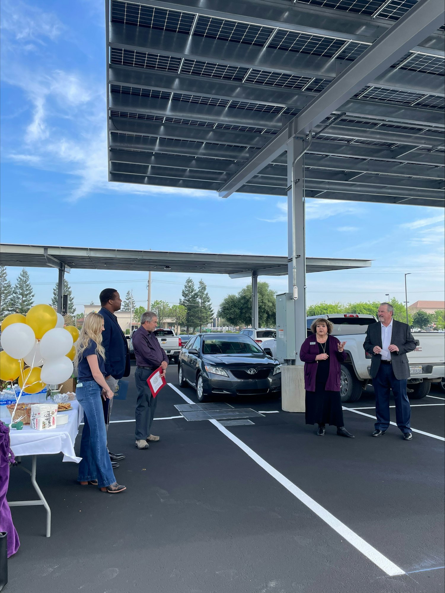 A group of people gathered in a parking lot with solar panels, balloons, and a refreshment table, likely at an event.