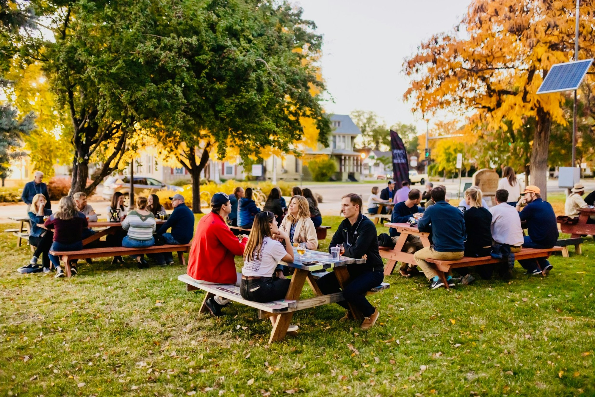 People socializing at picnic tables in a park with autumn trees.