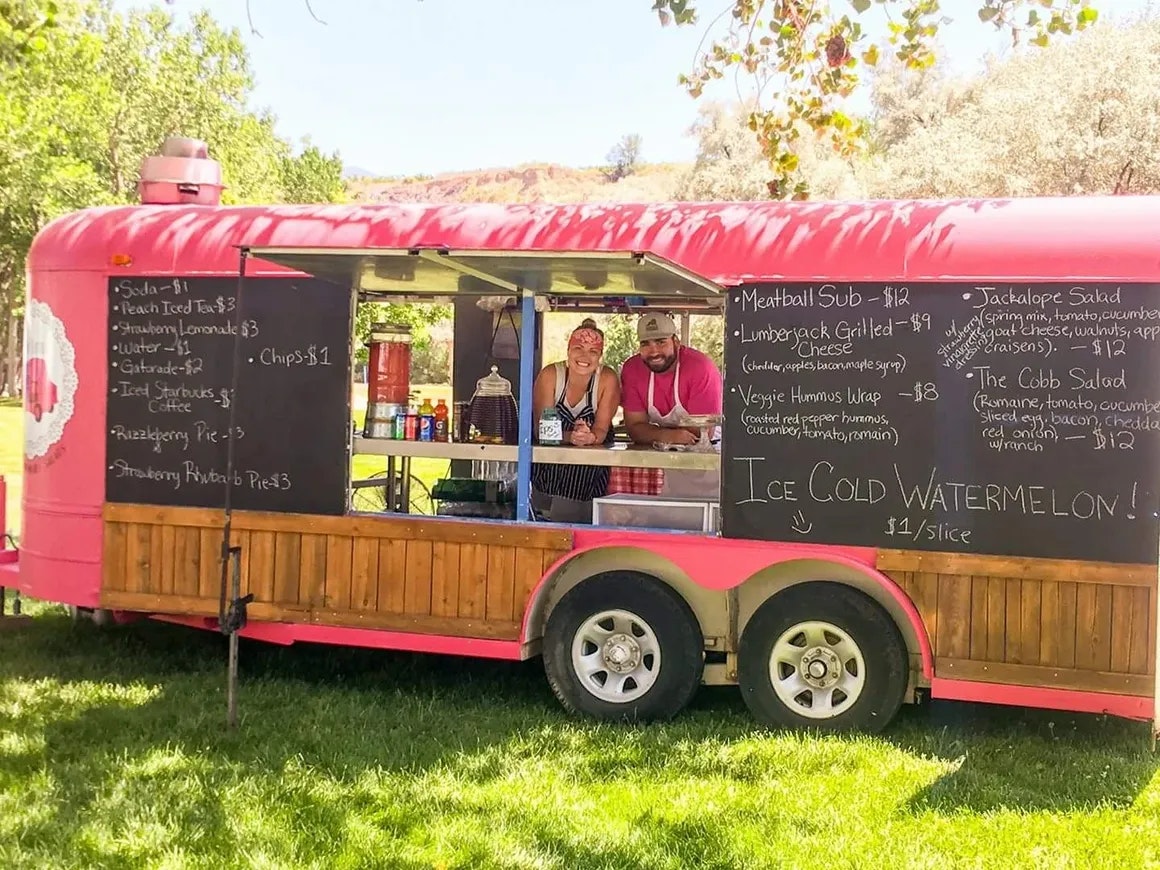 A red food trailer with an open service window and a menu board, surrounded by greenery, with two people inside.