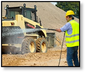 A construction worker in a yellow vest and helmet is power washing a vehicle at a construction site.