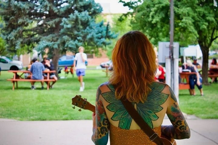 A woman with tattoos holding a guitar, facing a park with people sitting at picnic tables.