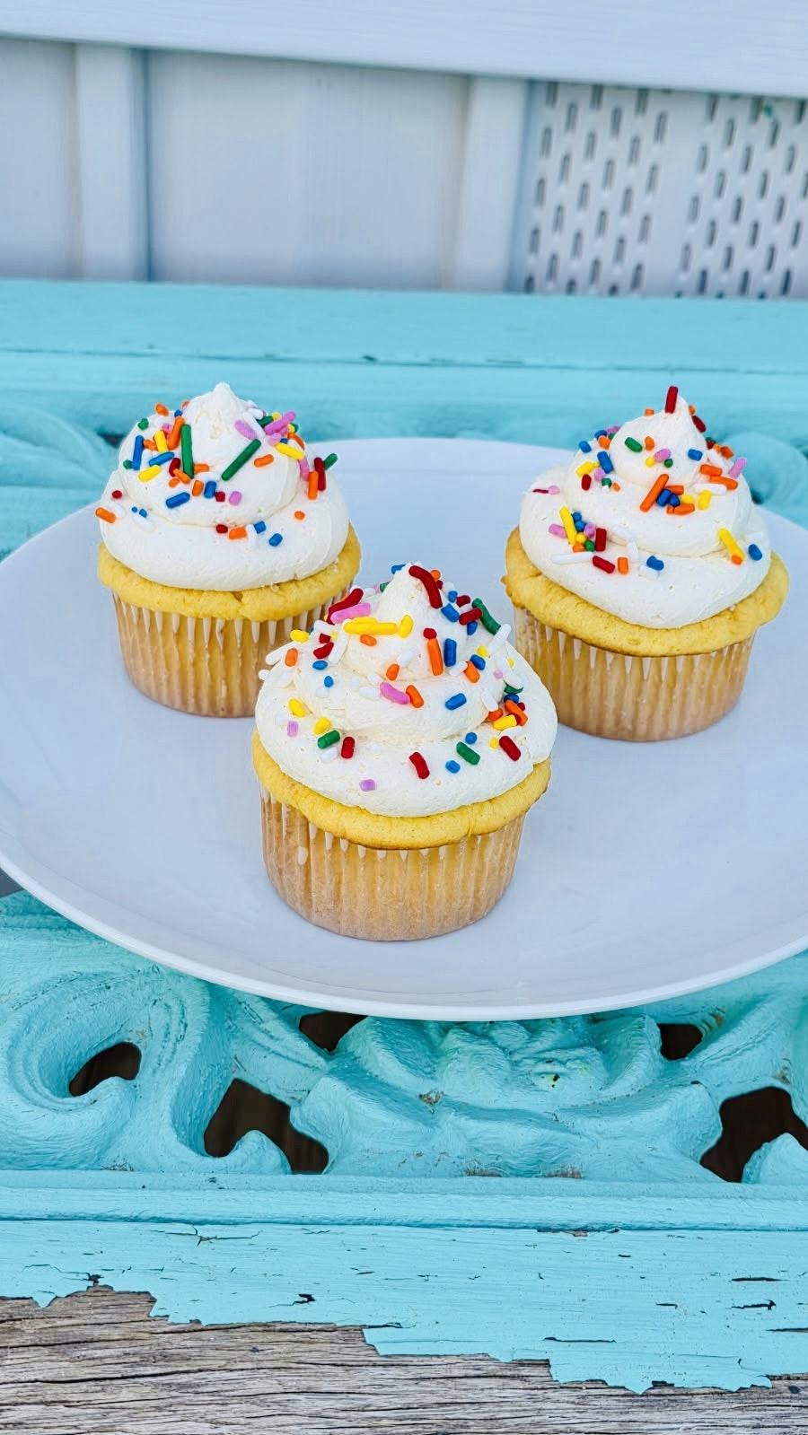 Three colorful cupcakes with frosting and sprinkles on a white plate, set against a turquoise background.