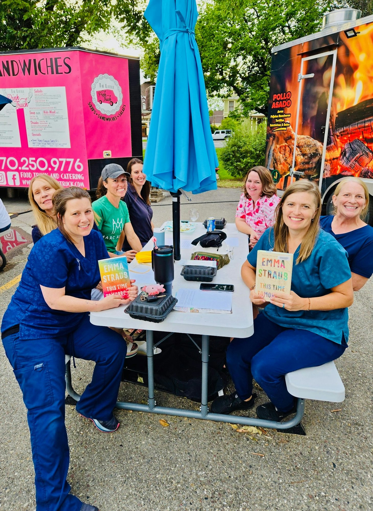 A group of women sit at a picnic table by food trucks, enjoying food and holding books, under a blue umbrella.
