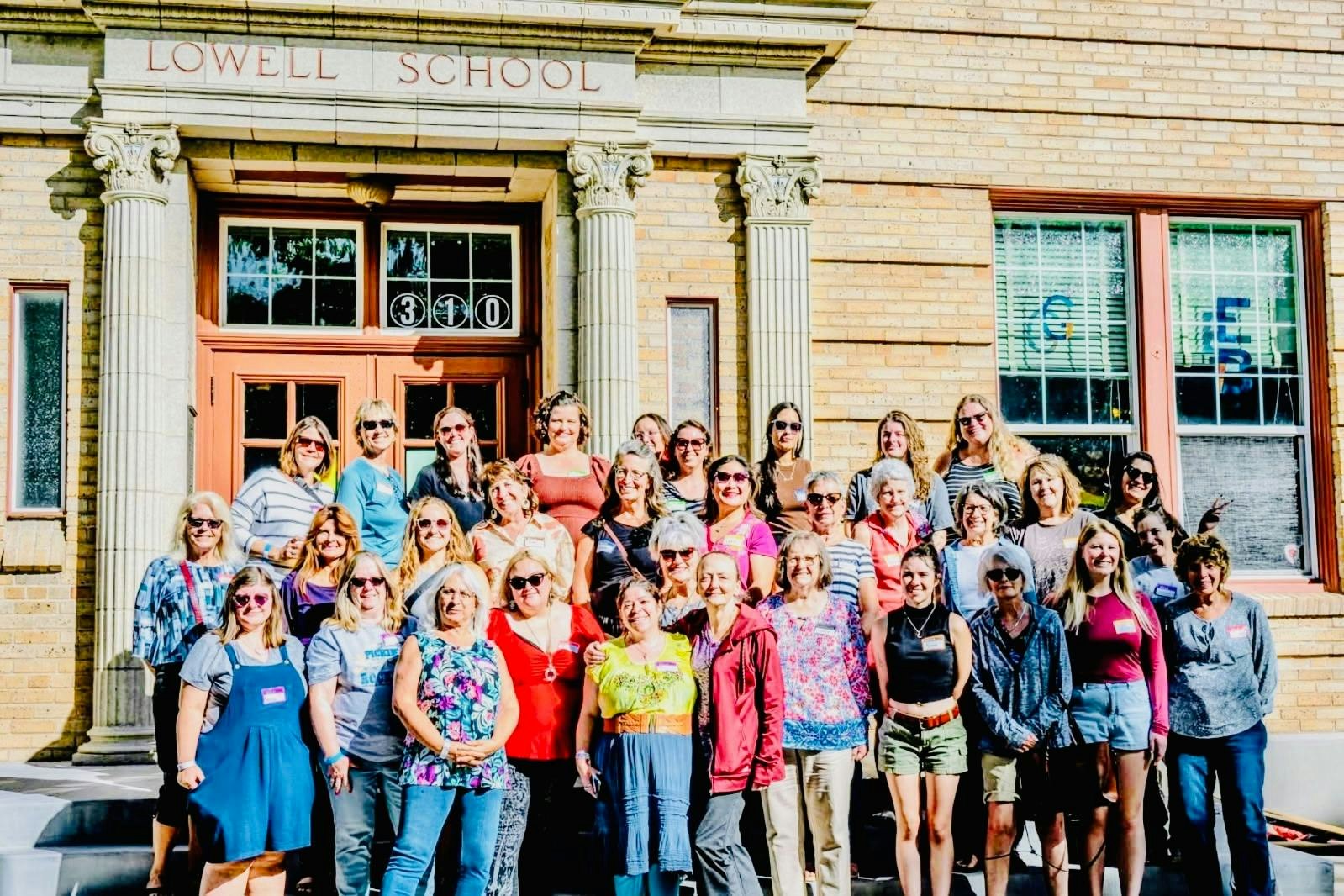 A large group of people, mostly women, poses in front of the Lowell School building on a sunny day.