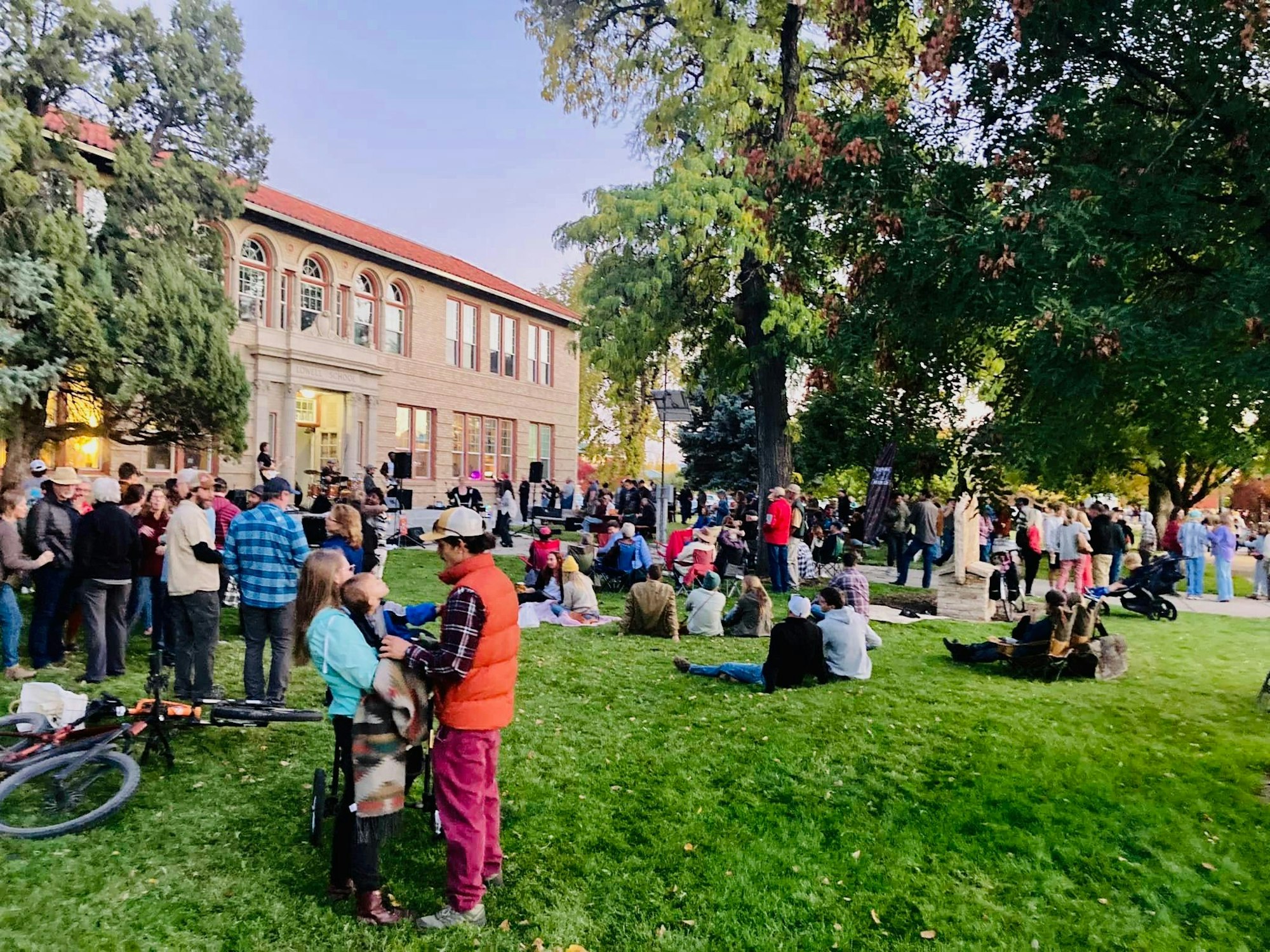 A crowd gathers outside a building in a grassy area, enjoying an event with people mingling, sitting, and live music.