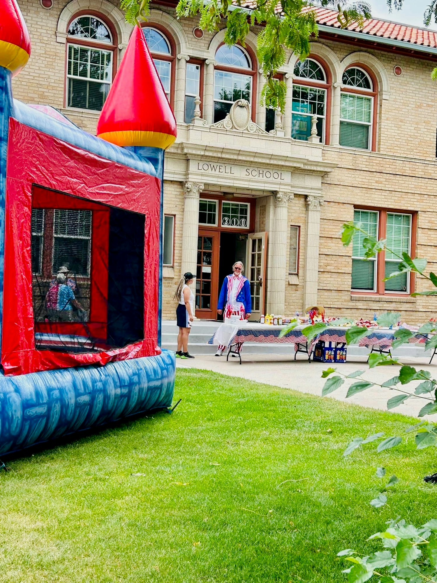 A school event with a bounce house, tables with food, and people socializing in front of a building labeled "Lowell School."