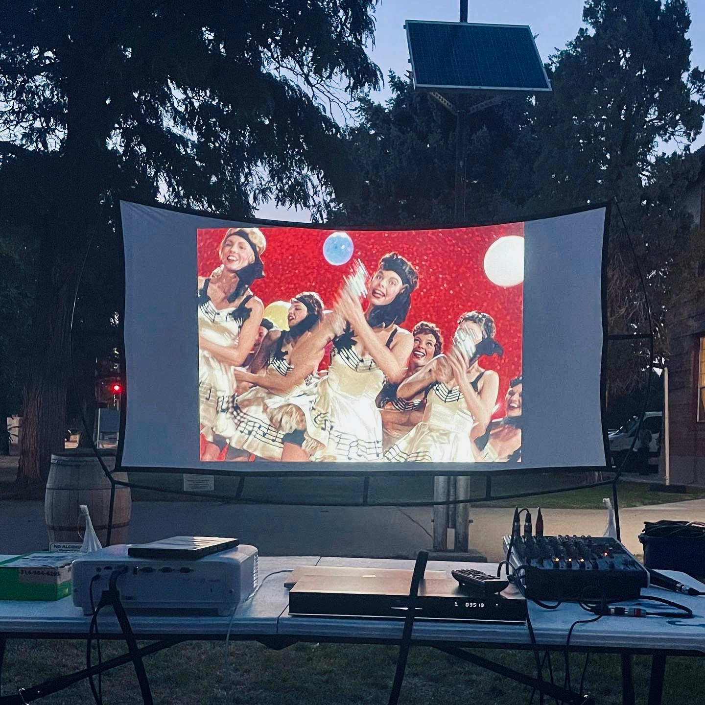 A projector screen displays a vibrant scene with performers in costumes, celebrating with balloons against a red background.