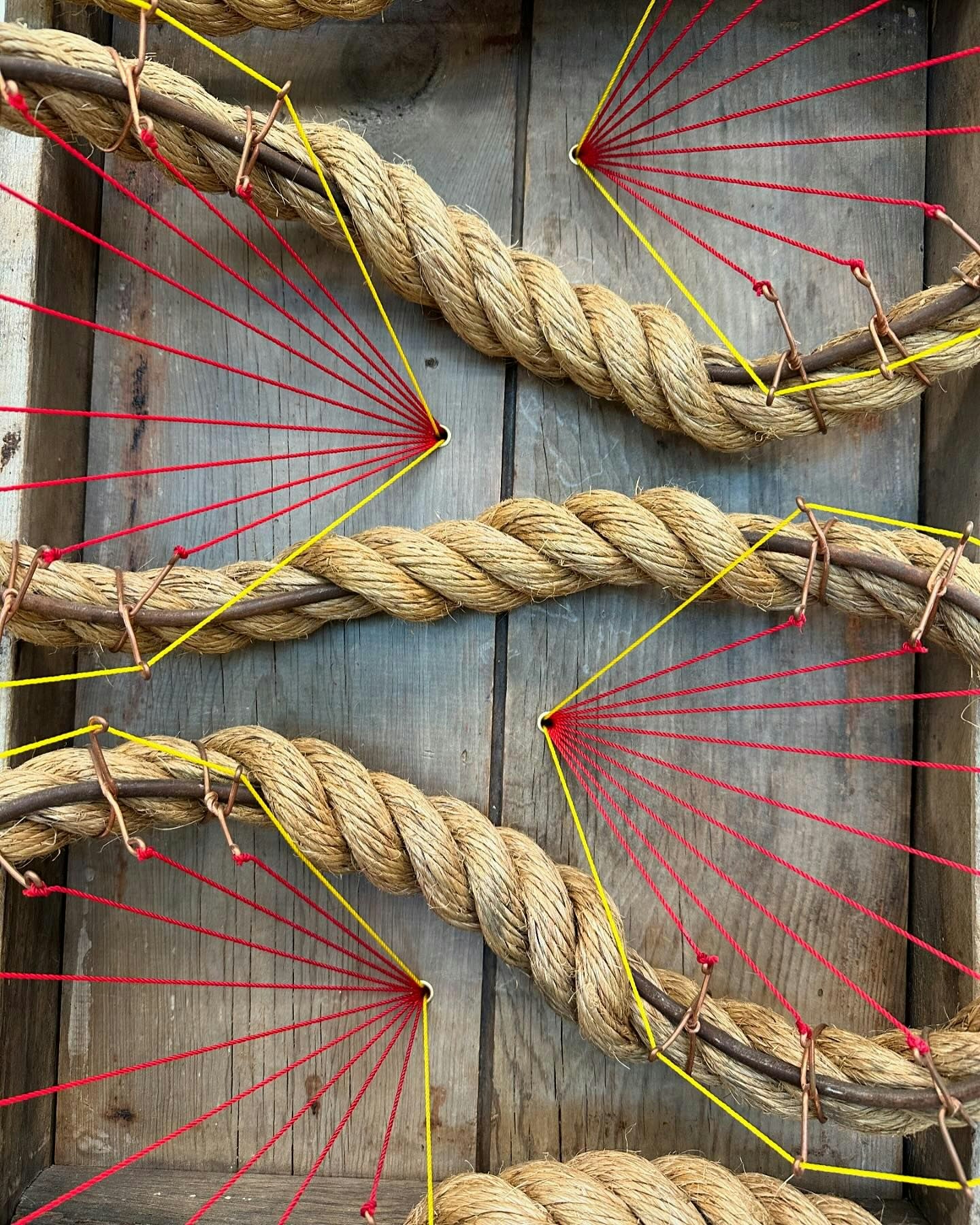 Thick ropes arranged in a hashtag pattern on wood, with red and yellow threads creating radial designs anchored at their intersections.