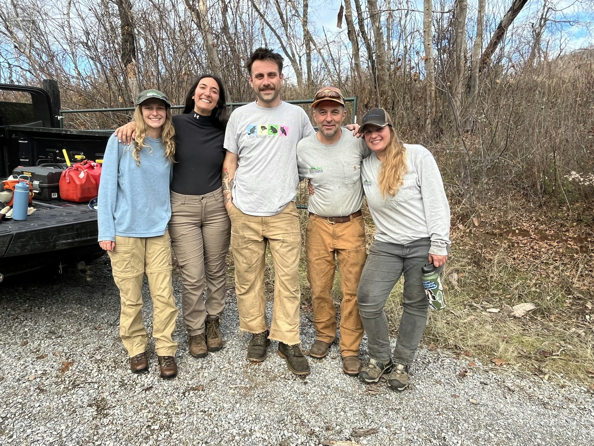 Five people smiling, standing on a gravel area near a truck, with trees in the background. Casual outdoor attire.