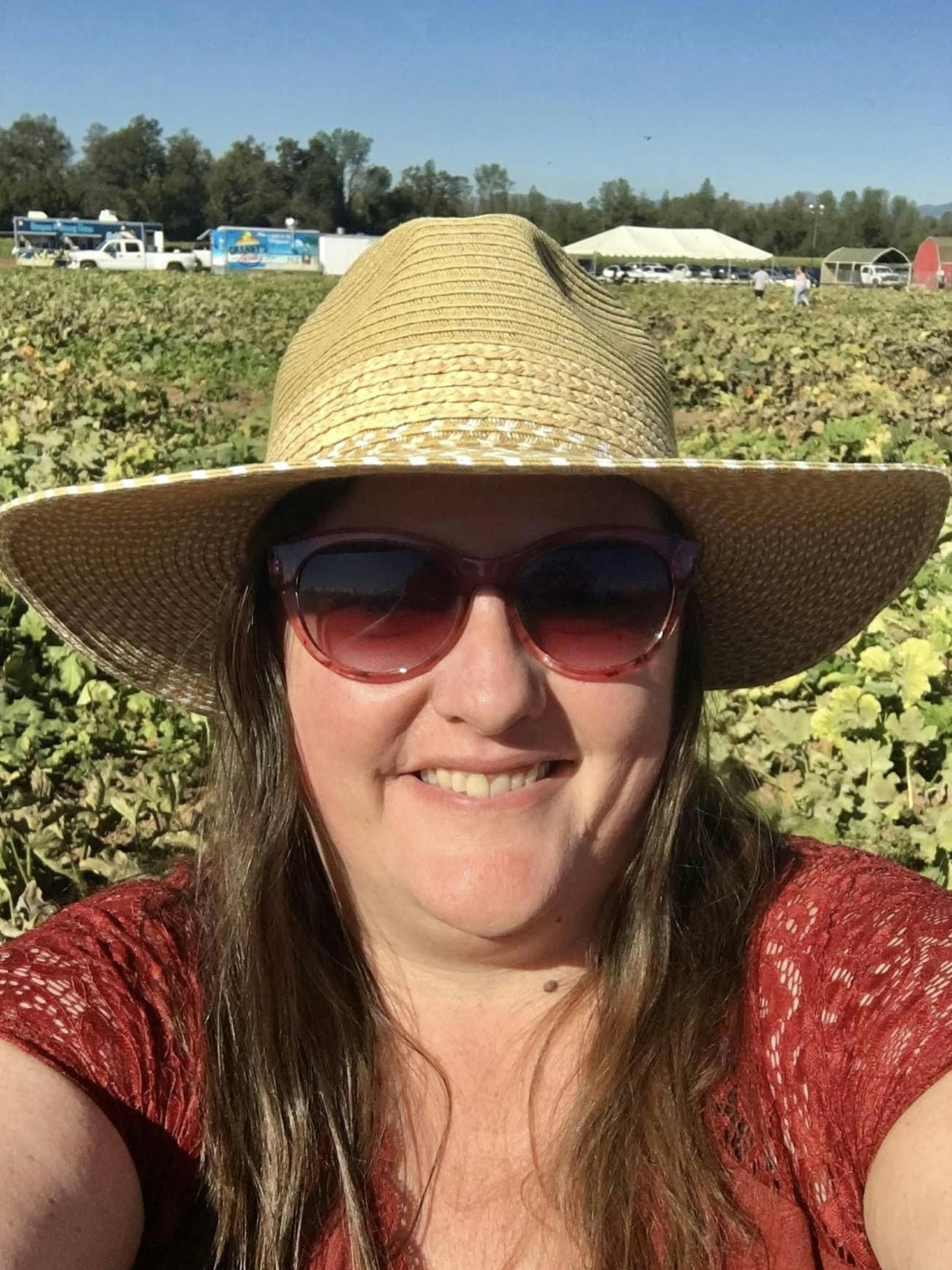 A person with sunglasses and a sun hat smiling in a field with tents and vehicles in the background.