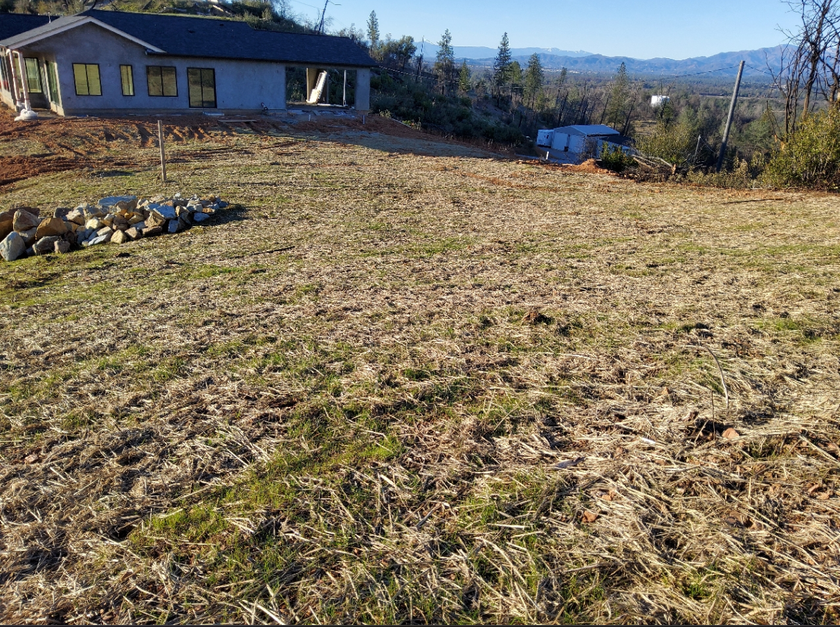 A house under construction with a patchy grassy field, rocks, and a mountainous forest background.