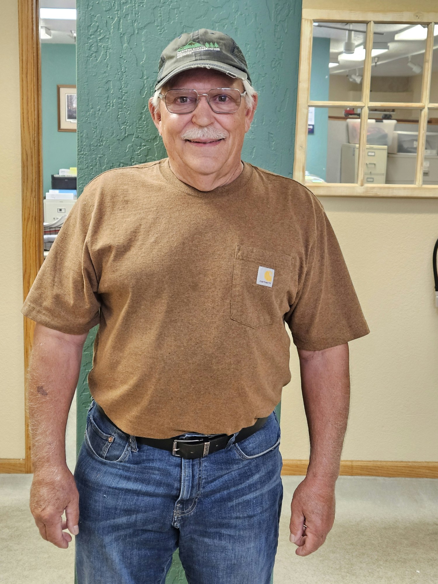 An older adult wearing glasses, a cap, a brown shirt, and blue jeans stands inside a room with green and beige walls.
