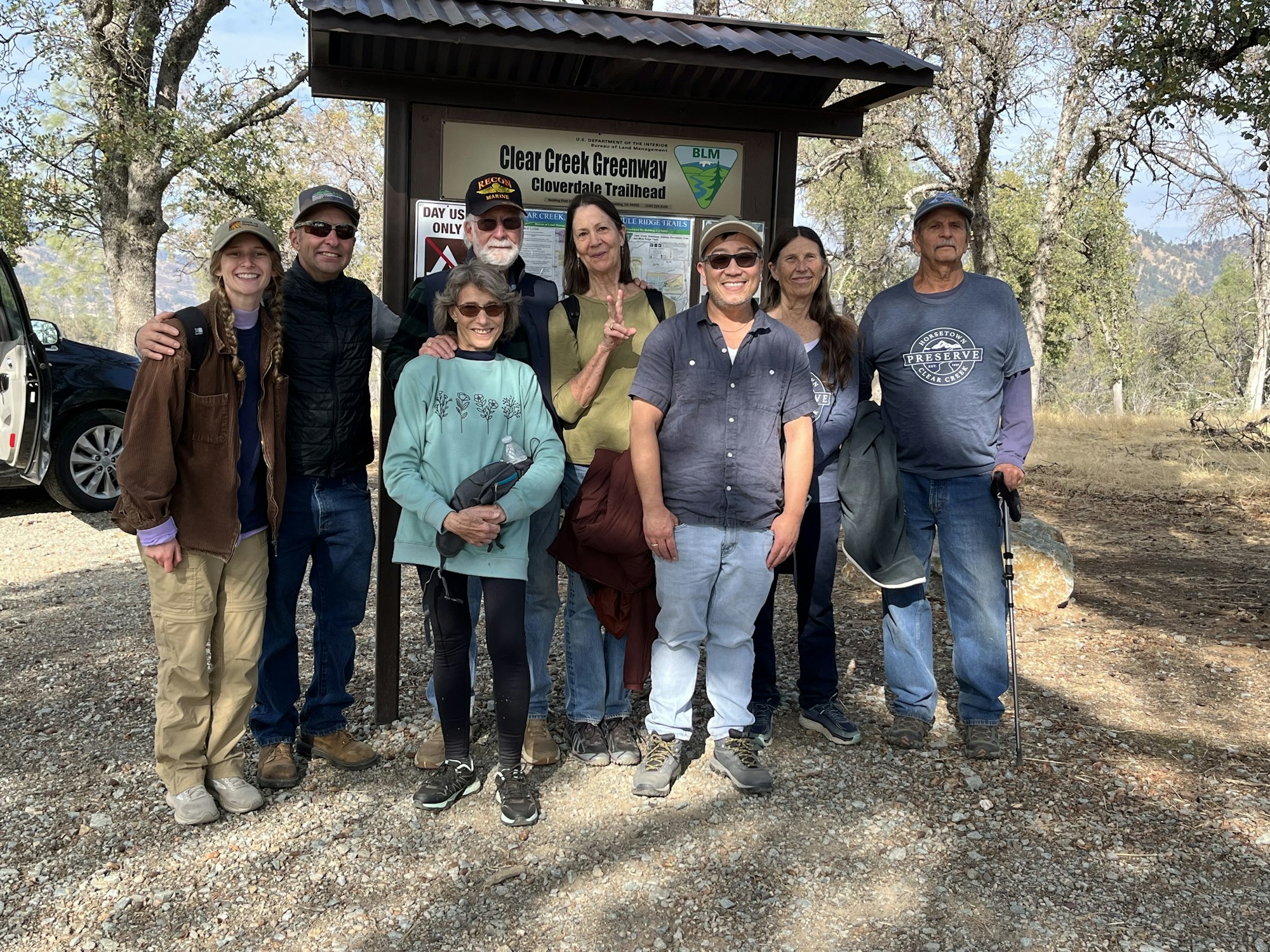 A group of people posing in front of a trailhead sign in a wooded area.