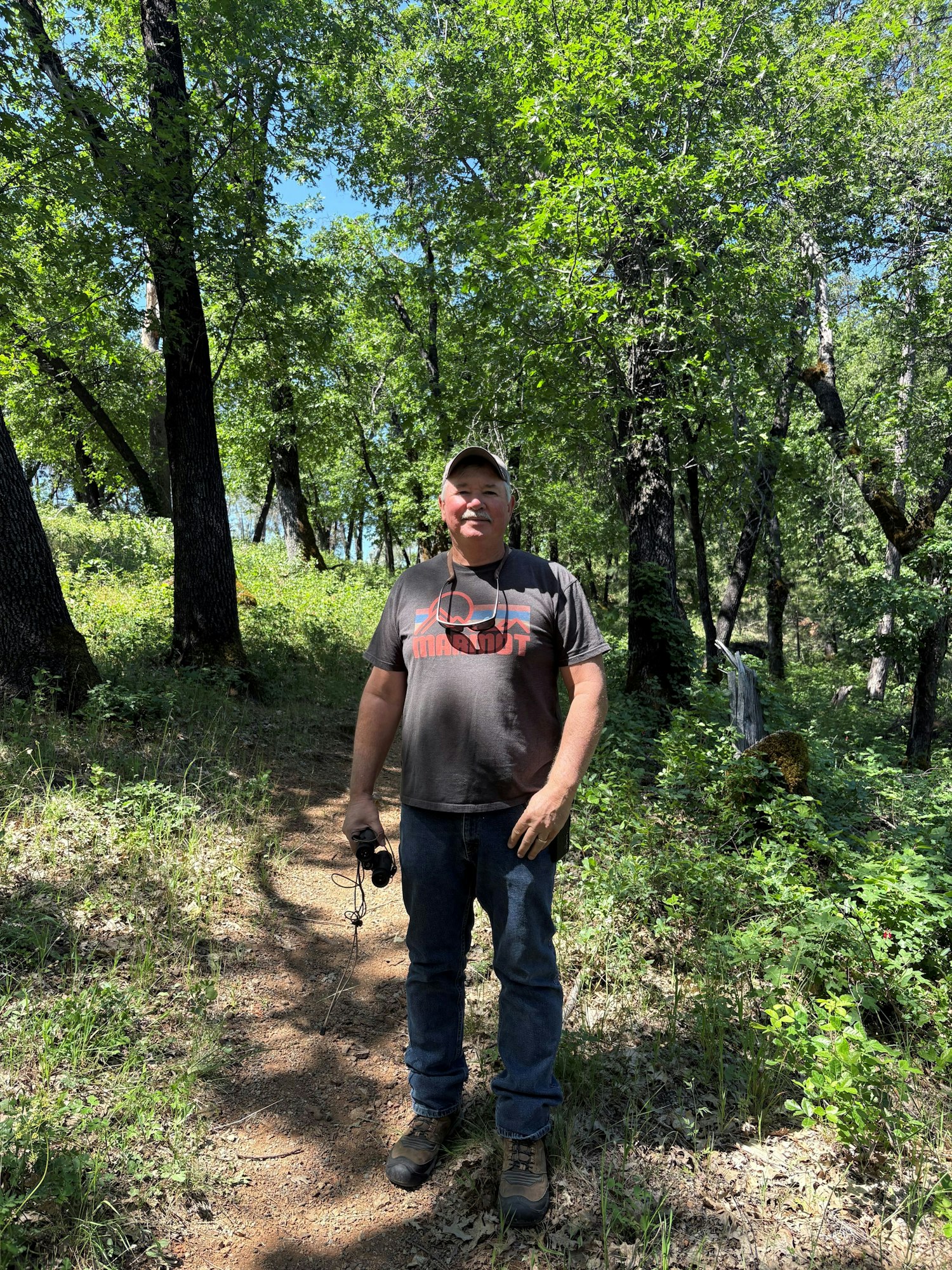 A person standing on a forest path, holding an object, with trees and greenery around.