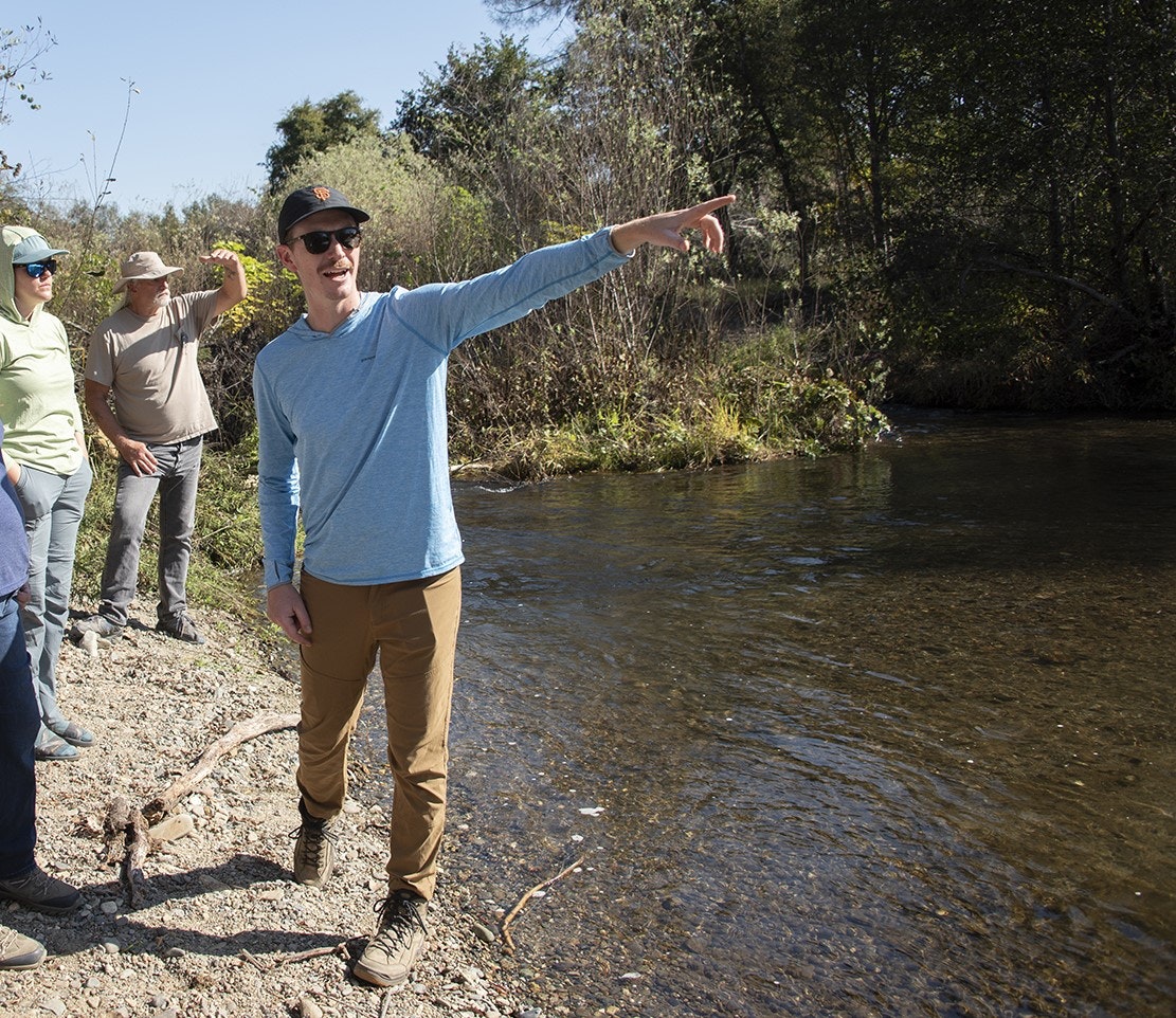 A group of people stands near a river; one person in the foreground points across the water.