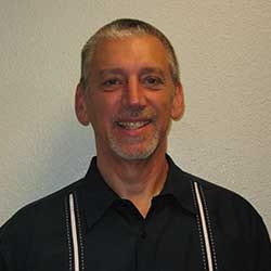 A man smiling, wearing a black shirt with white stripes, against a pale background.
