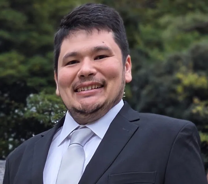 A person is smiling in a suit and tie, with greenery in the background. The image conveys a friendly and formal vibe.