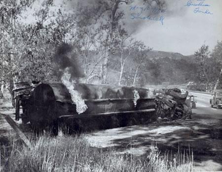 A black-and-white photo of an overturned, smoking vehicle beside the road, possibly after an accident.