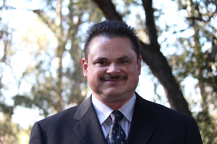 A person in a suit and tie is smiling, with trees in the blurred background.