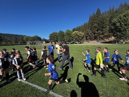 Children's soccer team on a field, exchanging post-match handshakes, surrounded by trees and blue sky.