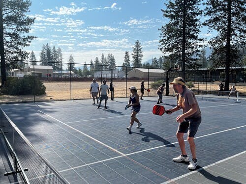 People playing pickleball on an outdoor court surrounded by trees under a blue sky.