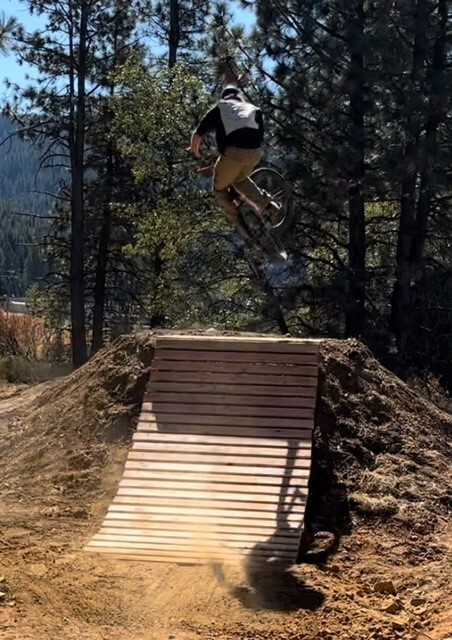 A person on a mountain bike jumps off a dirt ramp in a forested area with sunlight filtering through the trees.