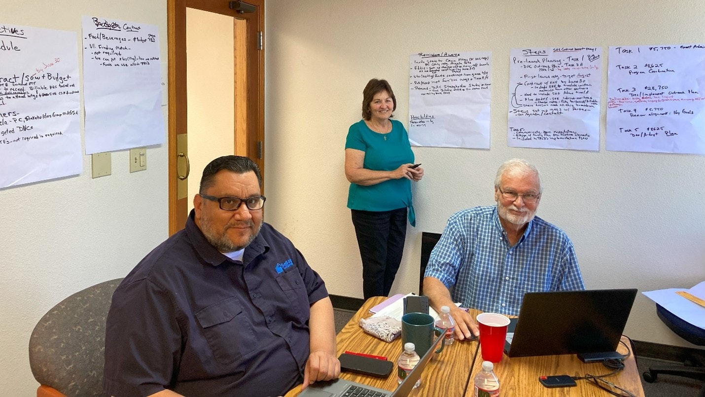 Three people in a meeting room with laptops, papers, and flip charts on the walls.