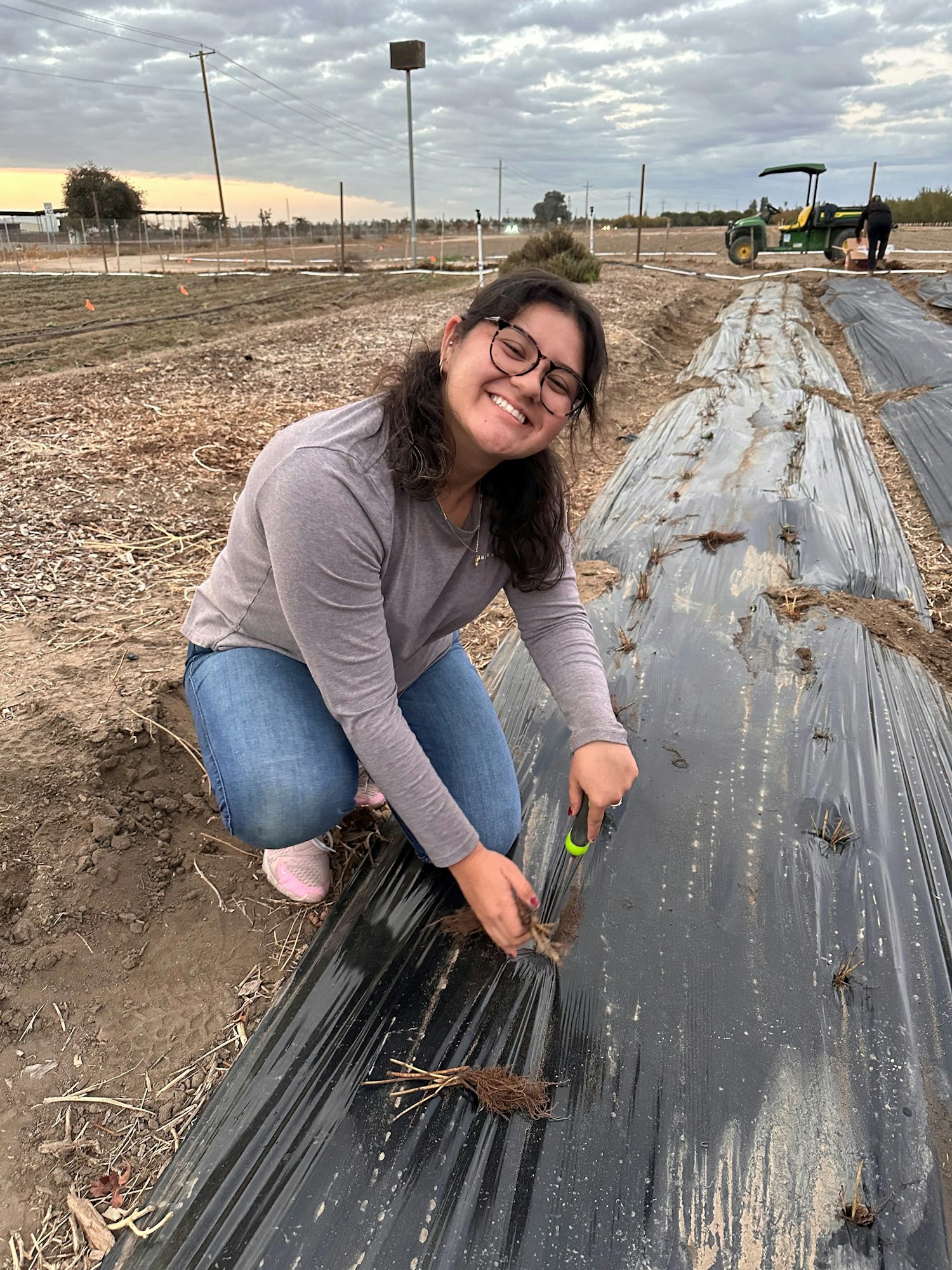 Jimena smiles while gardening, cutting plants under a cloudy sky. A tractor is visible in the background.